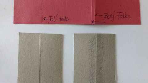 Zwei gefaltete Papiere in verschiedenen Farben liegen auf einem Tisch, während eine Anleitung zum Falten sichtbar ist.