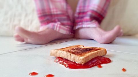 Ein Stück Toast liegt auf dem Boden, beschmiert mit Marmelade, während ein Kind in gestreiften Pyjamas barfuß sitzt.