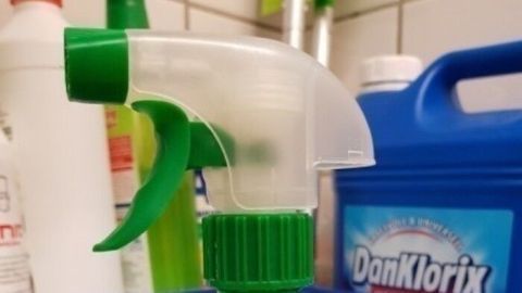 Eine grüne Sprühflasche steht auf einer Oberfläche, bereit, um Badreiniger selbst zu machen, im Hintergrunde Reinigungsmittel sichtbar.