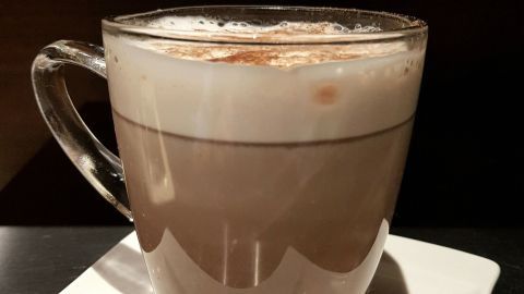 Eine cremige heiße Schokolade mit Zimt und Lebkuchengewürz steht in einem Glas auf einem weißen Teller.