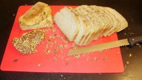 Ein frisch geschnittenes Brot mit Saaten liegt auf einem roten Schneidebrett, umgeben von Brotkrümeln und einem Messer.