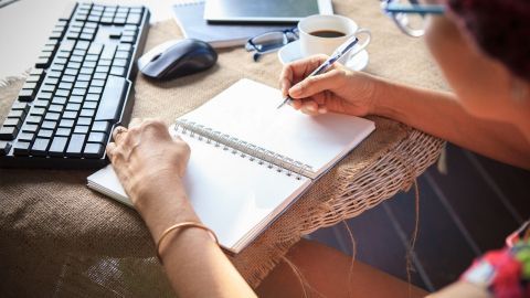 Eine Person schreibt in ein leeres Notizbuch am Tisch, um Ideen fürs Reimen und Dichten festzuhalten, umgeben von einem Kaffee.