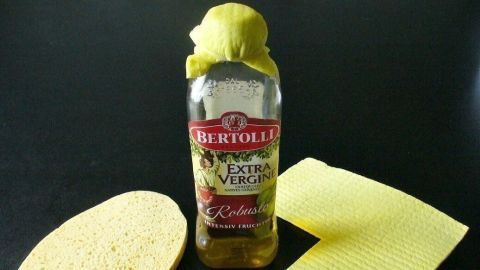 Eine Flasche Olivenöl steht neben einem Schwamm und einem Küchentuch, um Bratpfannen sparsam einzufetten.