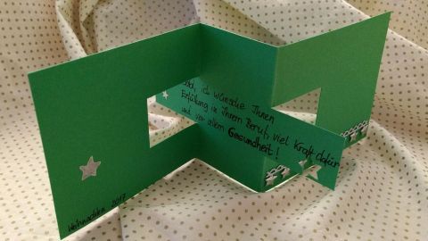 Eine aufgeschnittene Weihnachtskarte steht auf einer beige gestreiften Oberfläche, die in der Mitte grün ist und Sterne zeigt.