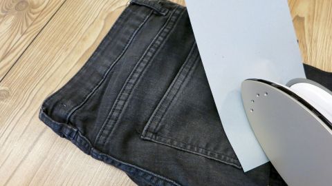 Auf einem Holzfußboden liegen dunkle Jeans mit einem Stück Löschpapier und einem Bügeleisen daneben, bereit zum Entfernen von Wachsflecken.