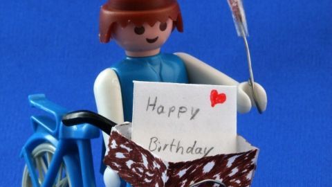 Eine Playmobilfigur fährt mit einem blauen Fahrrad, hält eine Glückwunschkarte mit "Happy Birthday" und eine rote Blume.