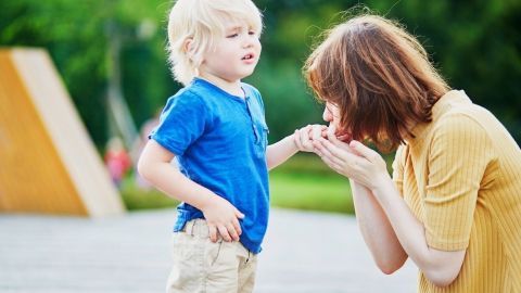 Ein Kind steht in einem Park vor einer Frau, die besorgt seine Hand hält und ihm Trost bietet.