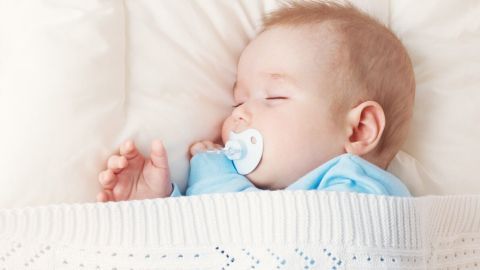 Wenn du dem Baby den Schnuller schmackhaft machen willst, kauf einen möglichst weichen und steck ihn dir idealerweise für ein Viertelstündchen in den Still-BH, in den an deinem Körper. Ein schlafendes Baby mit einem Schnuller liegt geborgen in einer Decke auf einem weichen Kissen, in ruhiger Umgebung.