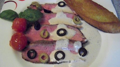 Zarte Scheiben von Vitello Tonnato mit einer cremigen Sauce, Tomaten, Oliven und einem knusprigen Brotstück auf dem Teller.