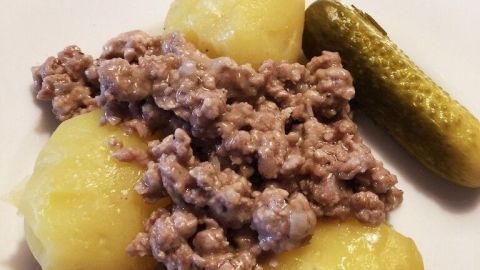 Ein Teller zeigt gekochte Kartoffeln, die mit einer würzigen Hackfleischsoße serviert werden, ergänzt durch eine Gewürzgurke.