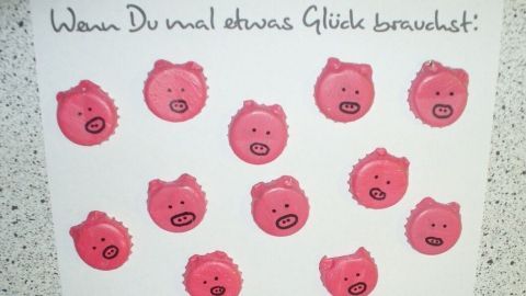 Auf einem weißen Blatt Papier sind viele kleine Glücksschweinchen aus Kronkorken abgebildet, die um einen Spruch gruppiert sind. Der Text lautet: "Wenn Du mal etwas Glück brauchst!"
