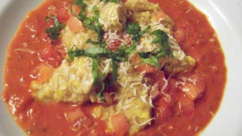 Kürbis-Malfatti liegen in einer tomatensaucenartigen Umgebung, garniert mit frischen Kräutern und Käse.