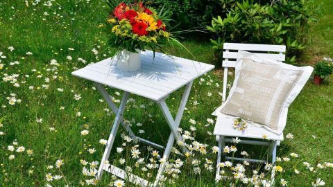 Ein weißer Tisch und Stuhl stehen im Gras, umgeben von Blumen, eine Vase mit bunten Blumen auf dem Tisch.