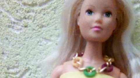 Eine Barbie-Puppe trägt eine selbstgebastelte Halskette und ein Armband, posiert in gelbem Kleid vor einer strukturierten Wand.