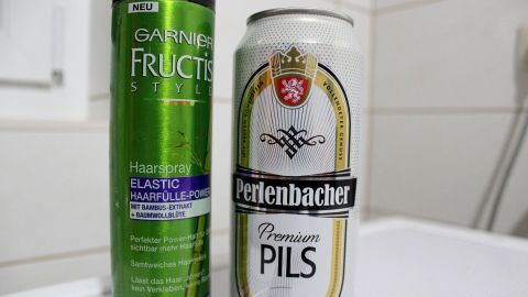 Eine Haarspray-Dose steht neben einer Bierdose, beide sind in einem Badezimmer auf einer weißen Ablage platziert.