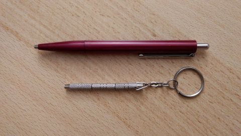 Ein roter Kugelschreiber liegt neben einem silbernen Schraubendreher für Brillen, der an einem Schlüsselring befestigt ist.