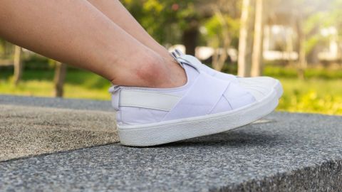 Ein weißer Turnschuh steht auf einer Betonfläche, während ein Fuß ihn leicht anhebt; im Hintergrund sind Bäume und Sonne sichtbar.