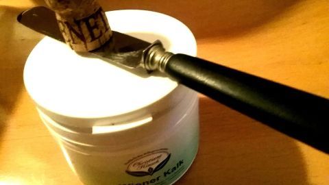 Eine Messerklinge liegt auf einem Behälter, während ein Korken darauf balanciert; der Kontext ist eine alltägliche Küchenszene.