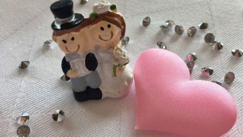 Eine Hochzeitsfigur steht neben einem pinken Herz, umgeben von glitzernden Dekorationen auf einem tischähnlichen Untergrund.
