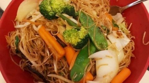In einer roten Schüssel liegen gebratene chinesische Nudeln mit buntem Gemüse, darunter Brokkoli und Karotten, die appetitlich arrangiert sind.