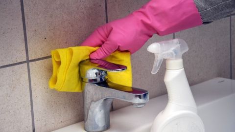Eine Hand mit pinken Handschuhen reinigt einen Wasserhahn mit einem gelben Tuch, während ein Sprühreiniger daneben steht.