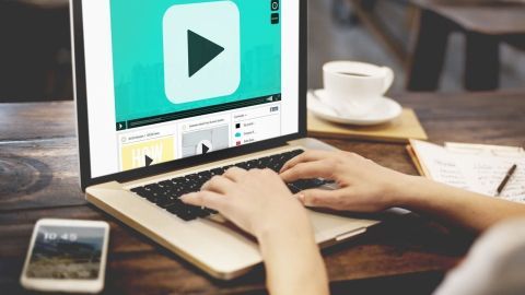 Eine Person sitzt am Tisch und spielt ein Video auf einem Laptop langsamer ab, während eine Tasse Kaffee daneben steht.