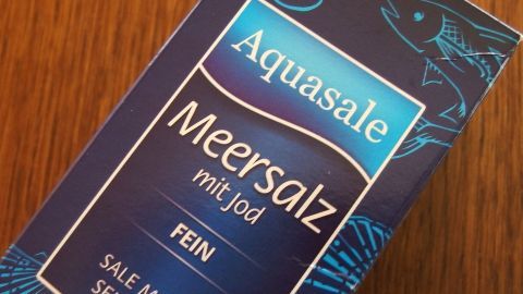 Meersalz kann effektiv gegen Pickel helfen. Mit seiner antibakteriellen Wirkung sorgt es für reinere Haut. Probier es als Gesichtswasser oder Peeling aus und erlebe die natürliche Kraft des Meersalzes. Eine blaue Verpackung von Aquasale Meersalz mit Jod liegt auf einem Holztisch. Text: "FEIN, SALE MARINO, SEL FIN" ist sichtbar.
