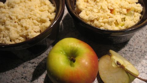 Ein frischer Apfel liegt neben zwei Schalen mit knusprigem Crumble, das Apfel und Nüsse enthält, auf einer dunklen Oberfläche.