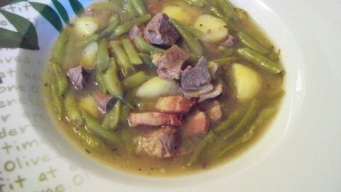 Ein herzhaftes Gericht, der Grüne-Bohnen-Eintopf mit Fleisch, in einer weißen Schüssel, umgeben von grünen Bohnen und Kartoffeln.