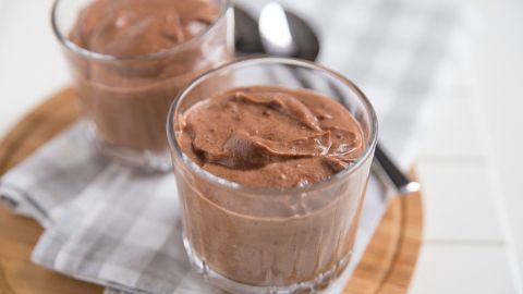 Zwei Gläser mit schokoladigem Dessert stehen auf einem Holzbrett, umgeben von einem gestreiften Tuch und Besteck.