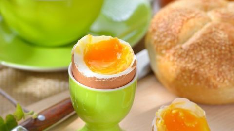 Ein perfekt gekochtes Frühstücksei steht in einem grünen Eierbecher, umgeben von Marmelade, Brötchen und einem Getränk.
