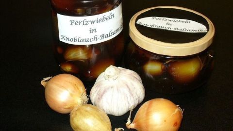 Glas mit Perlzwiebeln und Knoblauch-Balsamico-Essig steht neben frischen Zwiebeln und einer Knoblauchknolle auf einem Tisch.
