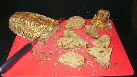Ein Brot liegt auf einem roten Schneidebrett, während es in unregelmäßige Scheiben geschnitten wird. Krümel verstreuen sich.