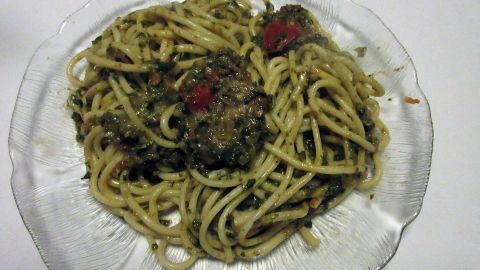Spaghetti auf einem klaren Glasteller, bedeckt mit einer Soße aus Spinat und Tomatenstücken auf weißem Hintergrund.