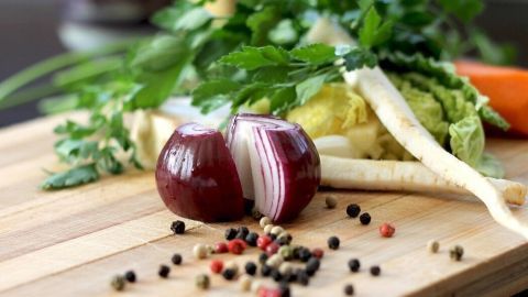 Eine rote Zwiebel ist halbiert auf einem Holzbrett, umgeben von frischem Gemüse und Gewürzen.