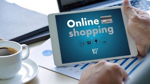 Ein Tablet zeigt "Online shopping" während jemand eine Tasse Kaffee in einem Büro mit Laptop und Dokumenten hält.