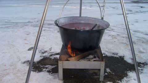 Ein großer, schwarzer Kessel steht über einem offenen Feuer auf einem Gestell im Schnee. Es wird gekocht.