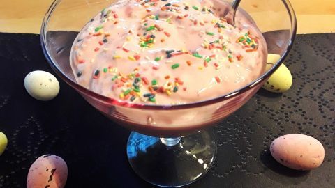 Eine Schüssel mit rosa Dessert, dekoriert mit Streuseln, steht auf einem schwarzen Tisch, umgeben von bunten Schokoladeneiern.