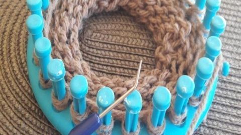 Ein Strickring in blauer Farbe zeigt eine braune Strickarbeit. Ein Häkelnadel wird eingesetzt, um das Stricken zu vervollständigen.