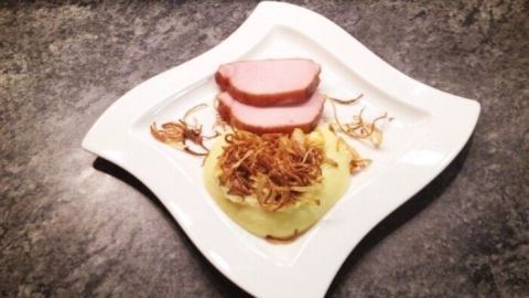 Saftiges Selchfleisch liegt auf cremigem Kartoffelpüree, umgeben von knusprigen Zwiebelringen, auf elegantem Teller.