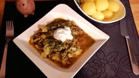 Eine Schüssel mit Schmorkohl und Hackfleisch, garniert mit Sauerrahm, auf einem Tisch mit Beilagen aus Kartoffeln.