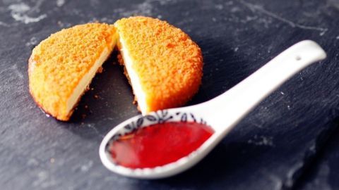 Frittierter Camembert liegt frisch auf einem schwarzen Teller, halbiert, neben einer Löffelportion süßer Sauce.