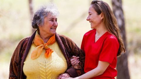 Eine ältere Frau wird von einer Pflegekraft unterstützt, während sie gemeinsam in einer natürlichen Umgebung spazieren gehen.