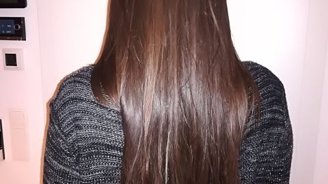Lange, seidige Haare fallen sanft herunter, während eine Person in einem dunklen Pulli vor einer hellen Wand steht.