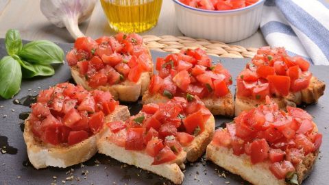 Getoastete Brotscheiben liegen mit frischen Tomaten belegt auf einer Platte, umgeben von Olivenöl, Knoblauch und Basilikum.