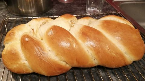 Ein goldbrauner Osterstriezel liegt auf einem Gitter, umgeben von einer dunklen Küchentheke und einem Glas.