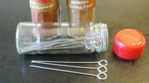Zwei Gewürzgläser stehen neben einer leeren Aufbewahrungsglas mit Rouladennadeln und einem roten Deckel auf einer dunklen Fläche.