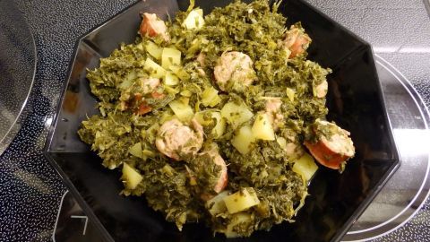 Eine Schüssel Grünkohl mit Mettwurst und Kartoffeln steht auf einem platten Herd, gut angerichtet und dampfend heiß.