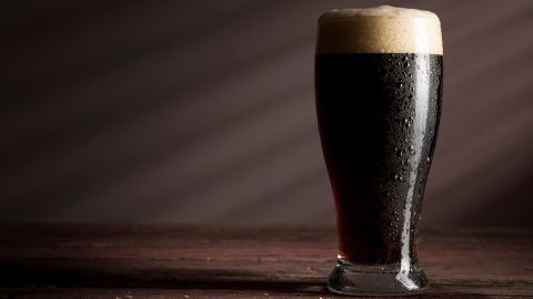 Ein mit Schaum gefülltes Glas dunkles Bier steht auf einem groben Holztisch, von sanftem Licht beleuchtet.