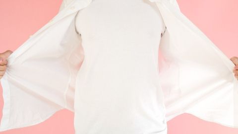 Ein weißes T-Shirt wird von einer Person mit ausgestreckten Armen präsentiert, vor einem pinken Hintergrund.